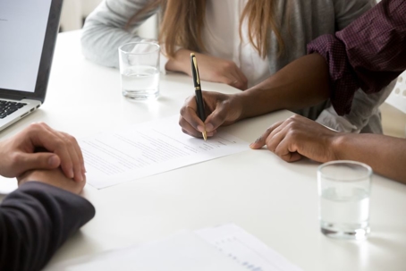 Man signing document on table with person sitting next to him.