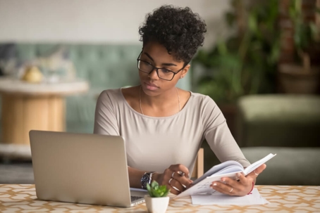 Woman with glasses on sitting at table with laptop and journal.