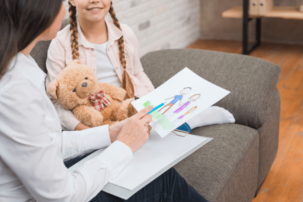 Woman sitting on a couch, holding a pencil and a child's drawing while talking to a young girl.